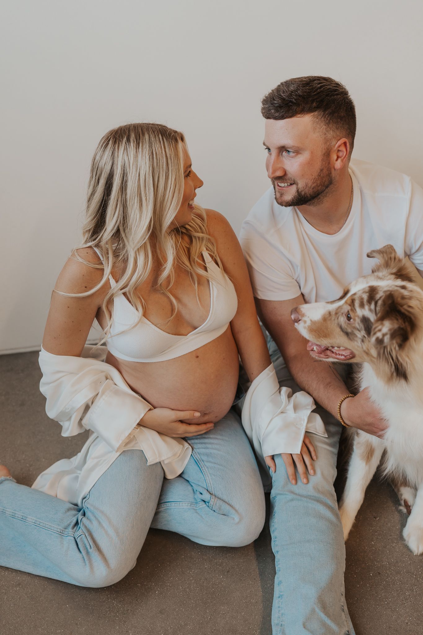 Expecting couple sits on floor with their Australian Shepherd dog in casual Michigan studio setting.