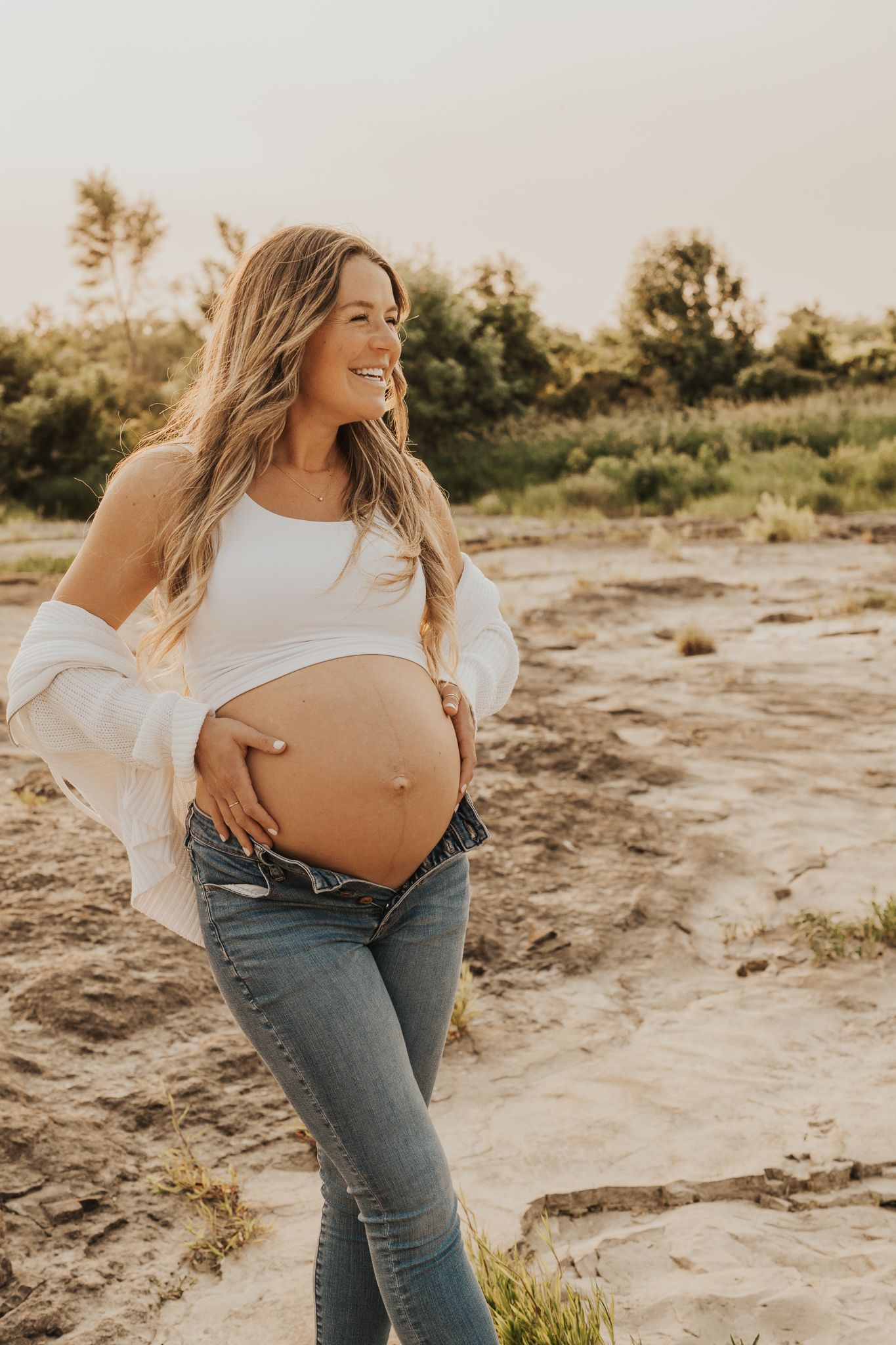 Expectant mother in jeans and white top poses outdoors showing baby bump.