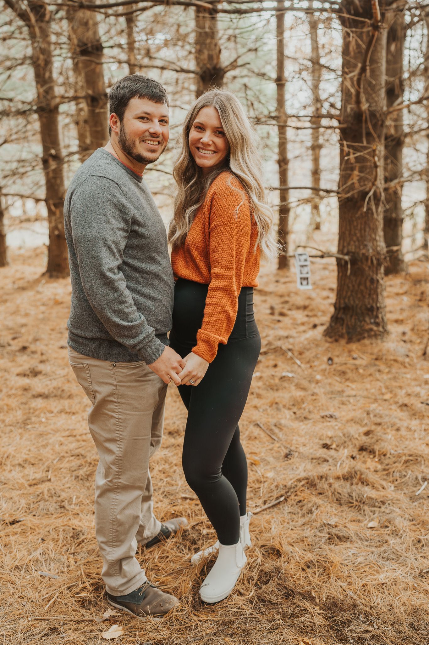 Couple in casual wear shares playful moment in pine forest during pregnancy announcement.