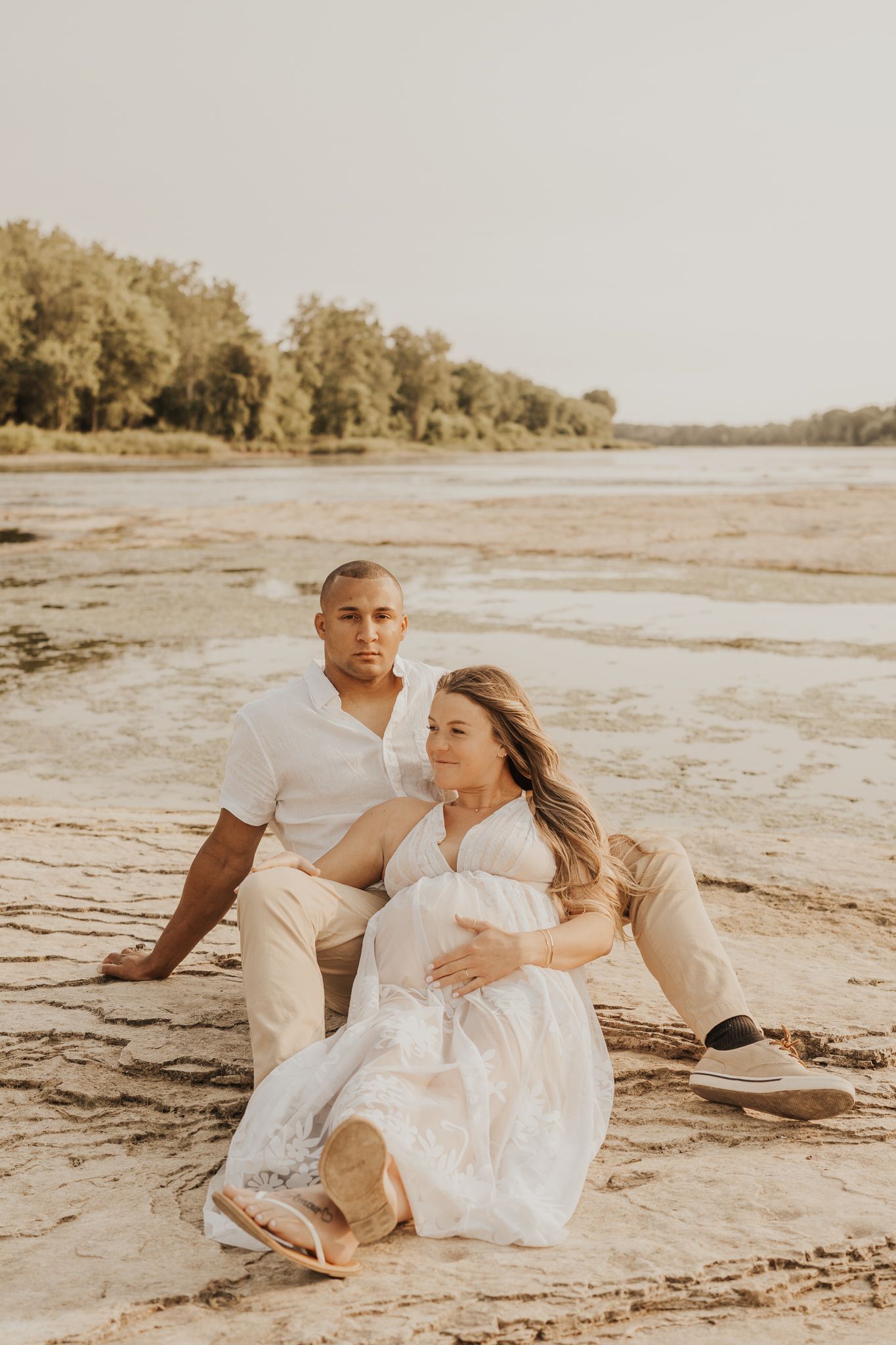 Couple relaxing on sandy riverbank during maternity photoshoot.