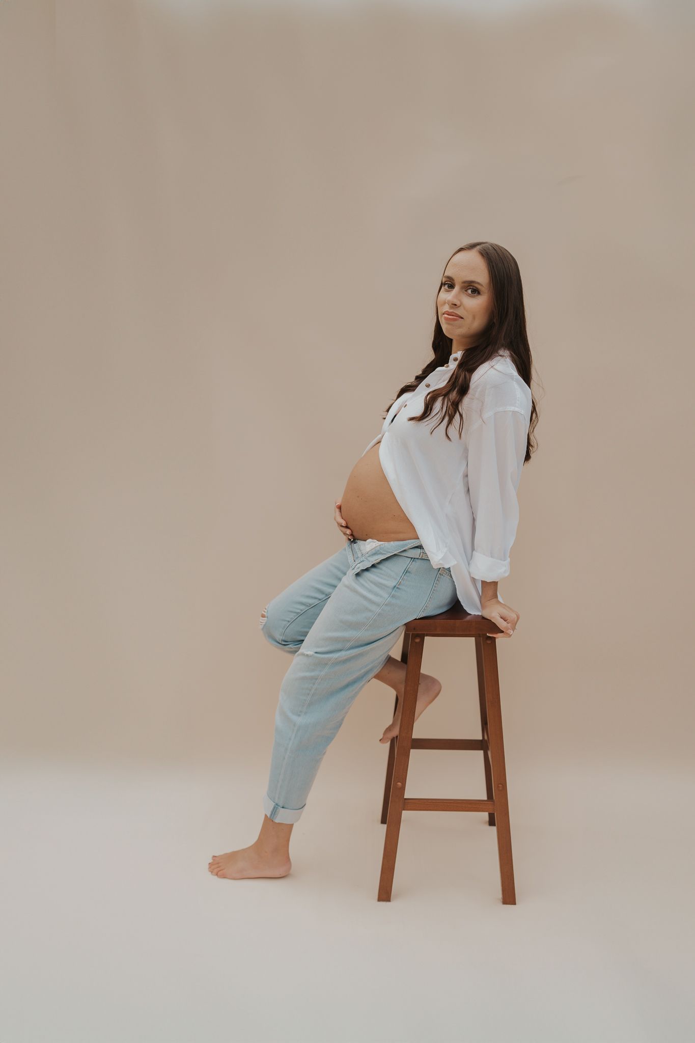 Maternity portrait of pregnant woman in white shirt and jeans sitting on wooden stool.