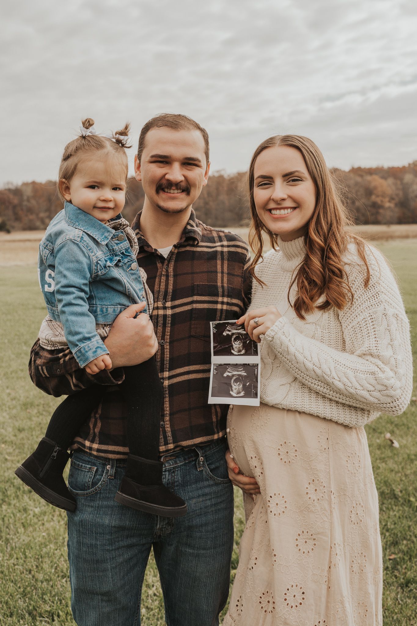 Couple in plaid and cream sweaters share pregnancy announcement with ultrasound photo in field.