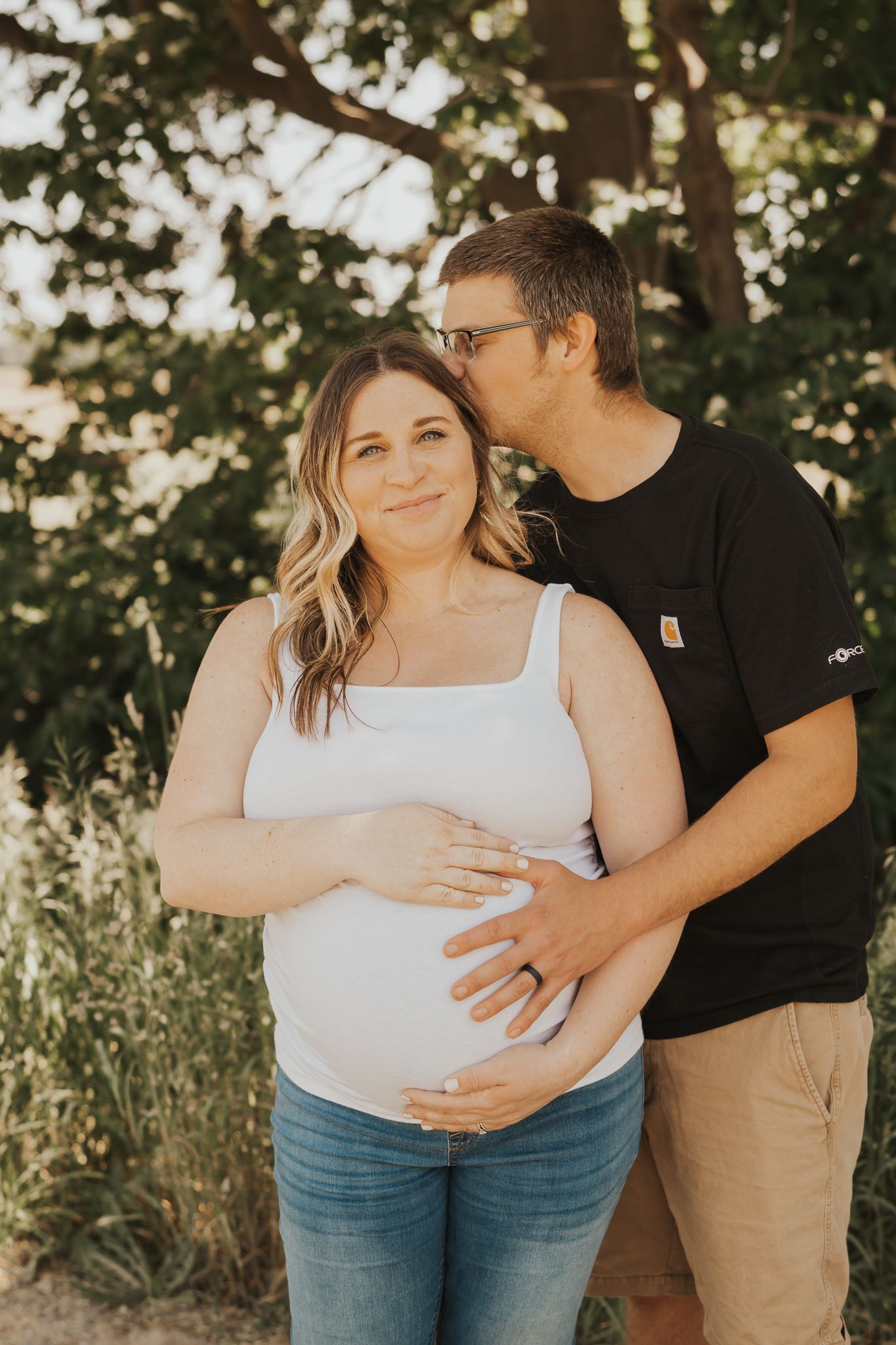 Couple shares tender moment during outdoor maternity photoshoot under trees.