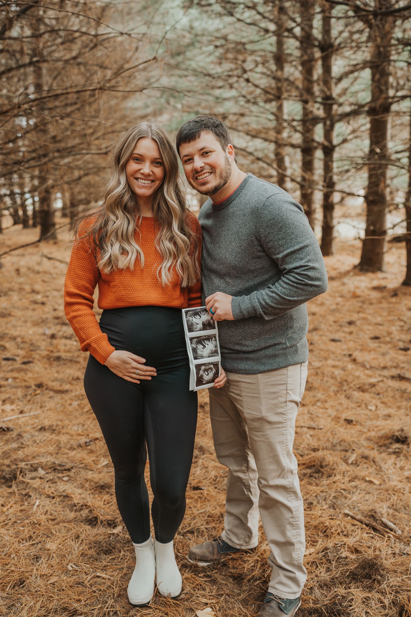 Expectant couple in orange and grey holding ultrasound photo in wooded setting in Michigan.