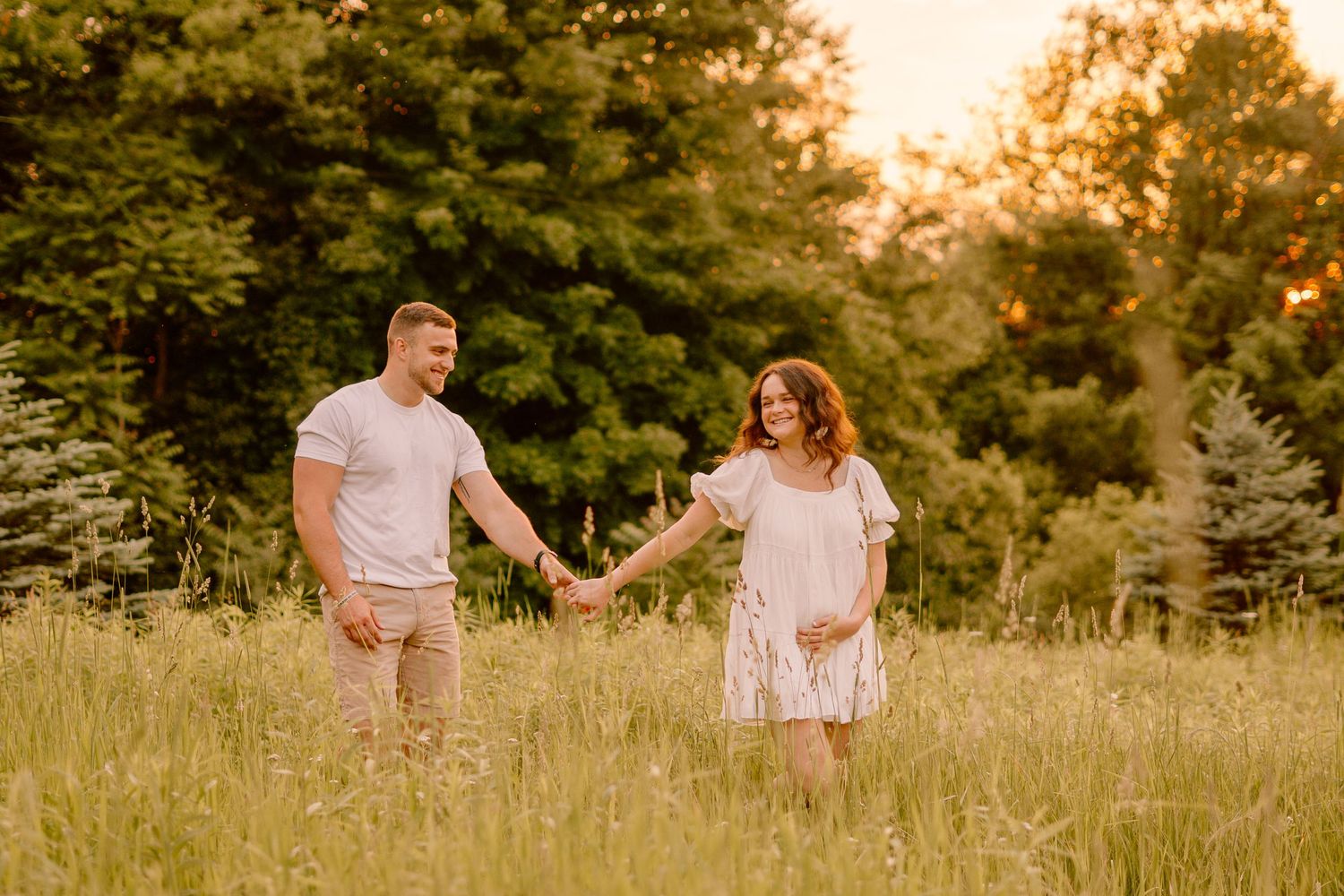 A couple holds hands while walking through a sunlit meadow at golden hour.