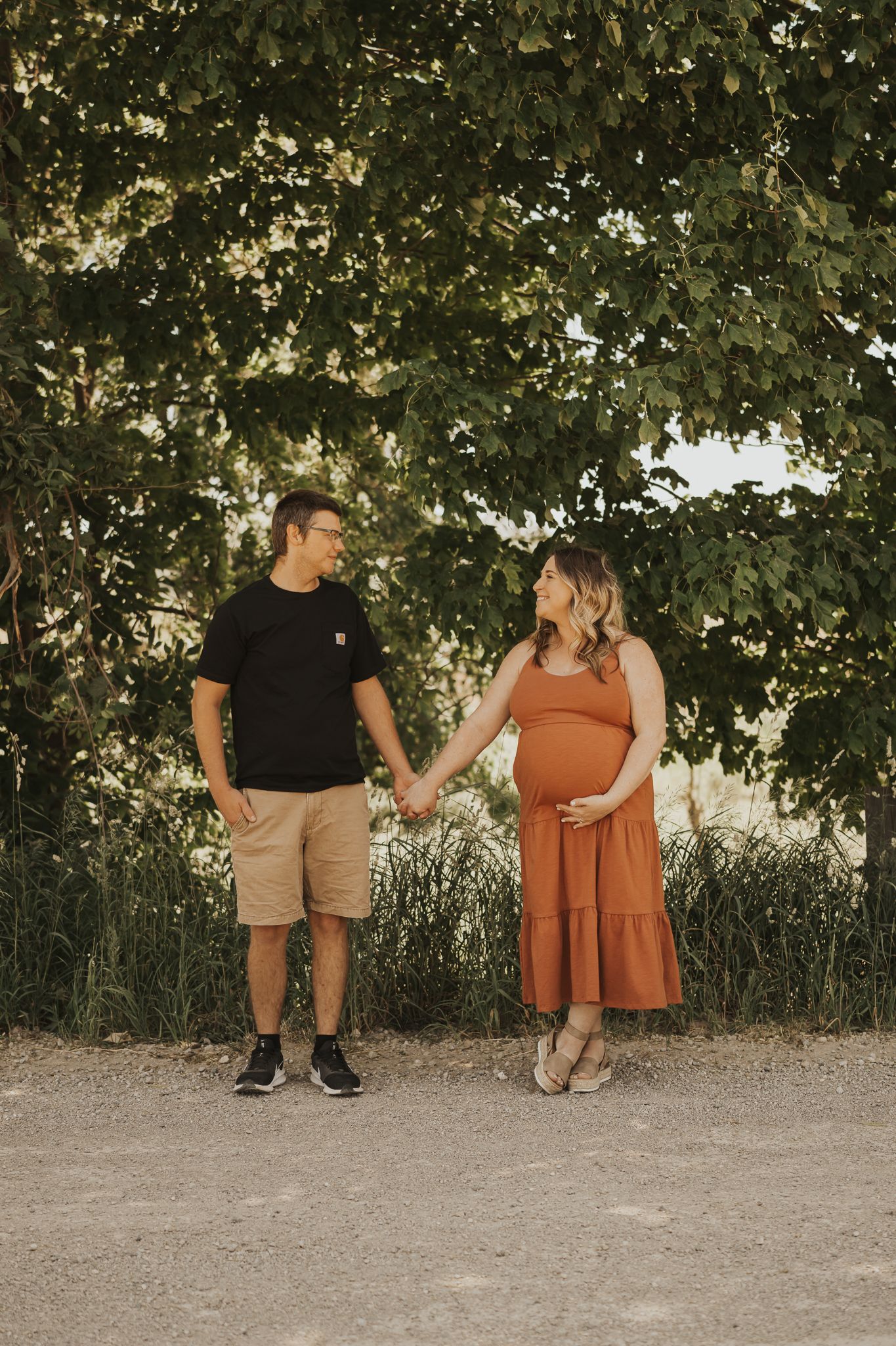 Couple sits under tree canopy during outdoor maternity photoshoot.
