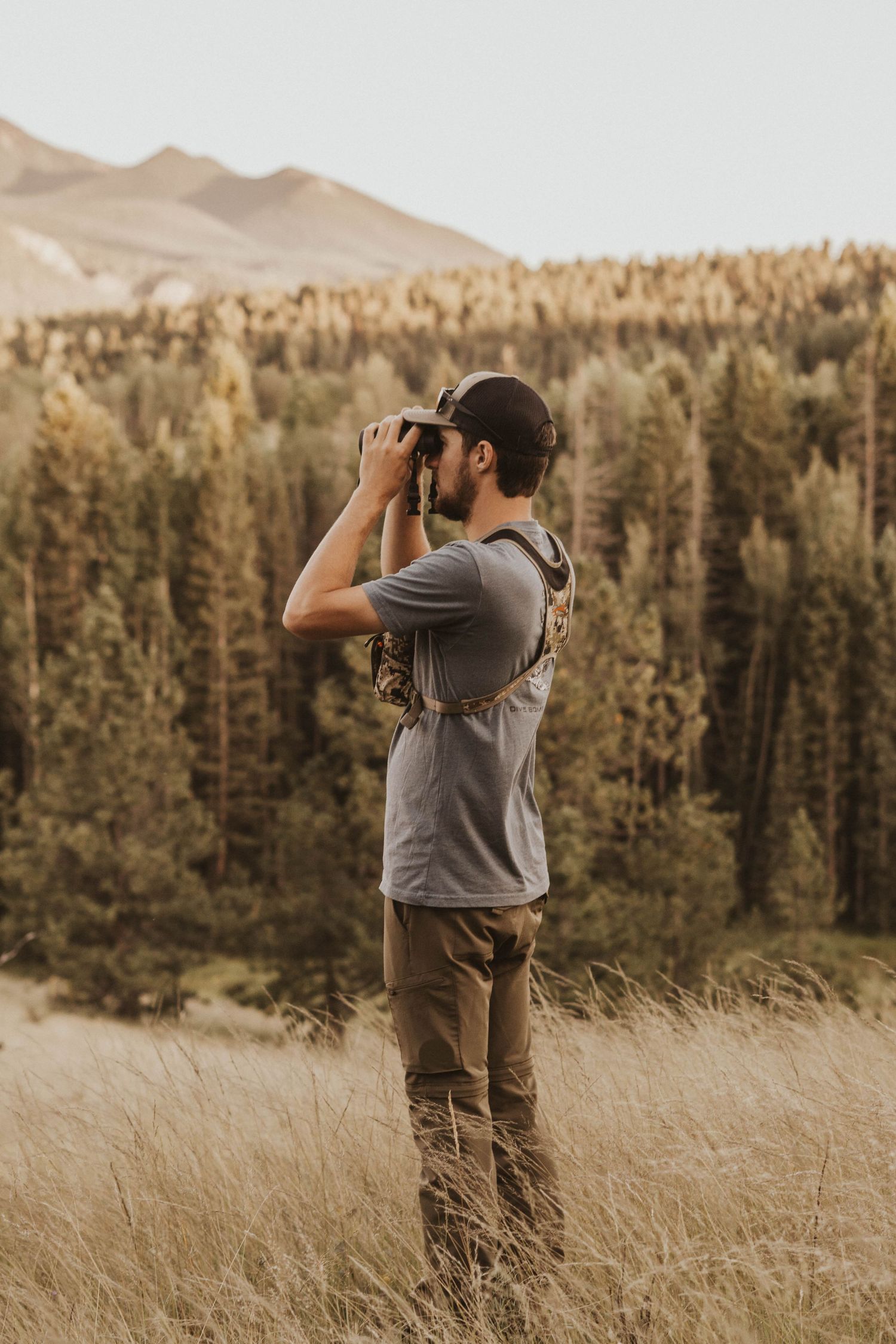 Person using binoculars to observe wildlife in a mountain meadow at sunset.