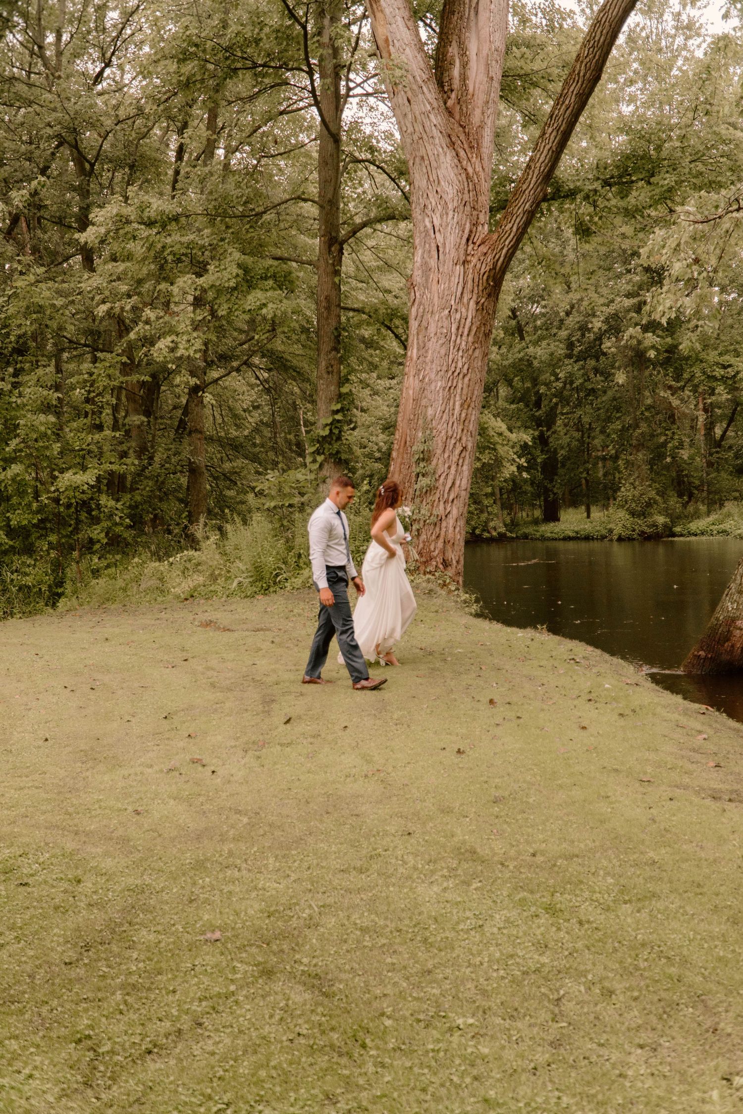 Couple walking together on a grassy hill under tall trees.