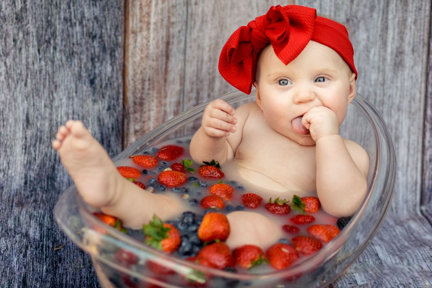 Baby girl wearing red bow sitting in glass bowl filled with strawberries and blueberries.