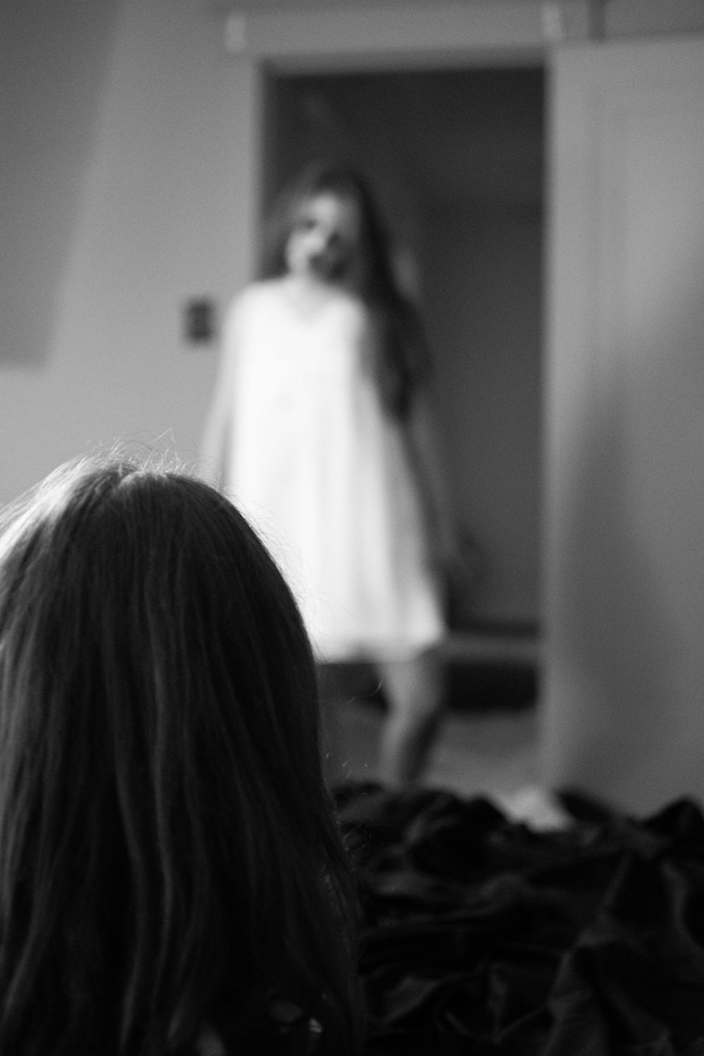A blurry black and white photo showing a figure in a white dress standing in a dark doorway.