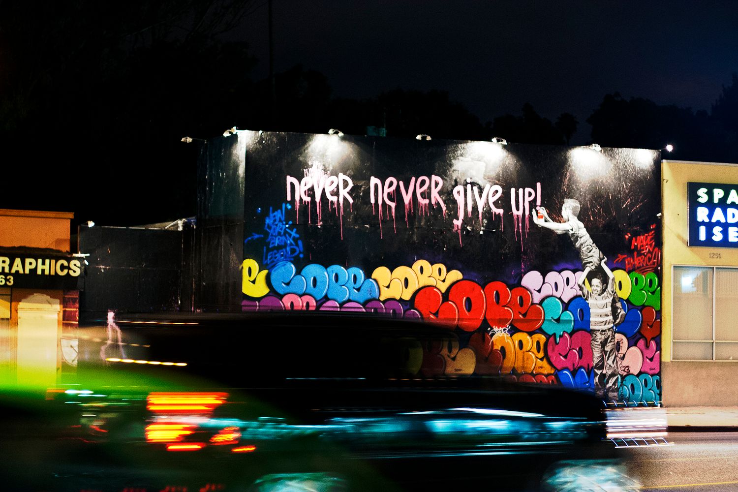 A graffiti wall at night displaying 'Never Never Give Up' in neon blue lighting.