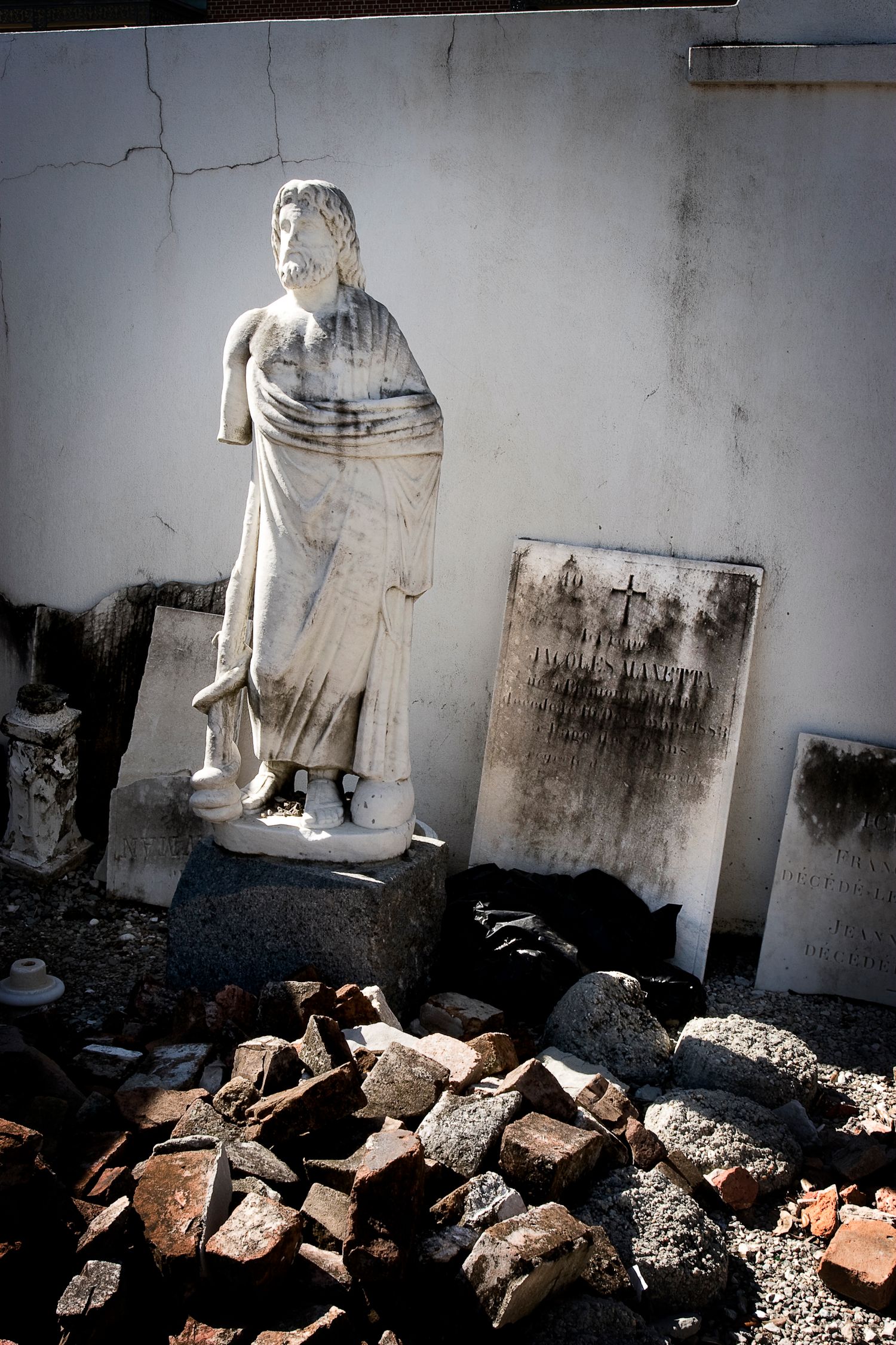 Weathered classical statue stands among broken stones and damaged gravestones.