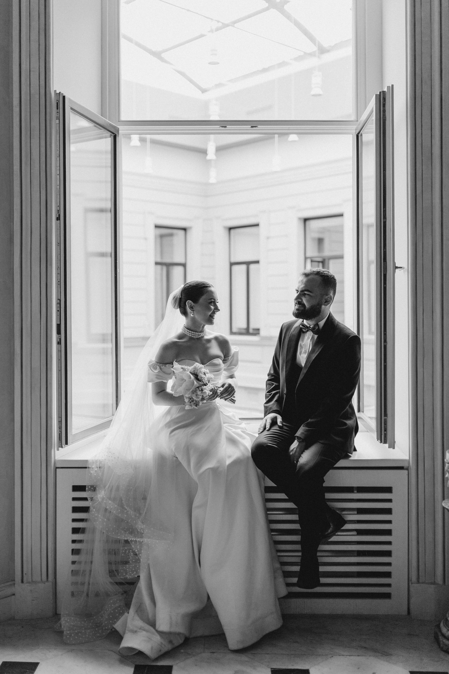 Black and white portrait of couple sitting on windowsill in elegant interior setting.