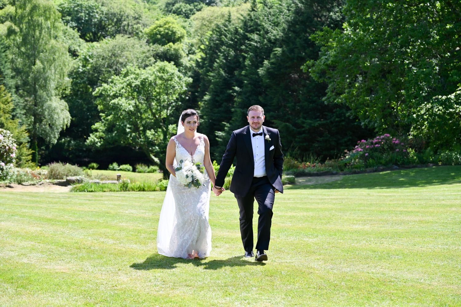 Newlyweds walking hand in hand across a sunny green lawn in formal wedding attire.