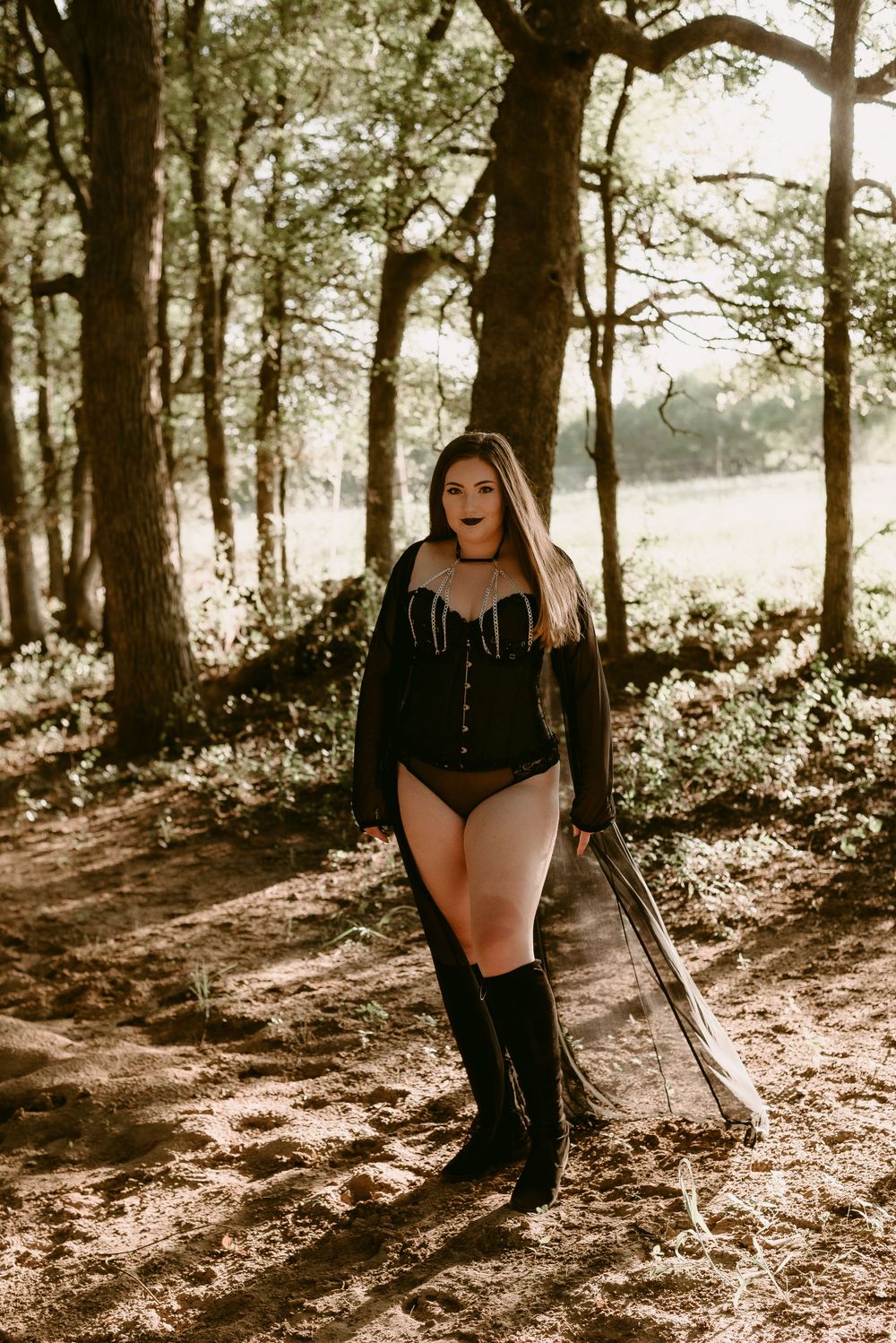 Forest path photoshoot featuring black corset and knee-high boots in filtered sunlight.