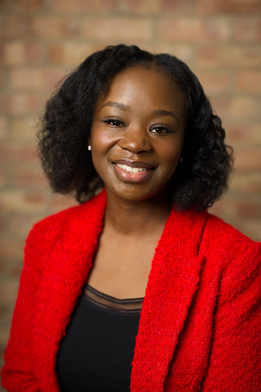 Professional headshot featuring bright red blazer against brick wall background.