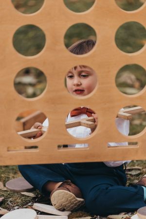 Child peeks playfully through circular holes in a wooden panel during a wedding.