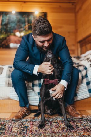Person in navy suit playing with black dog on patterned carpet in wooden cabin.