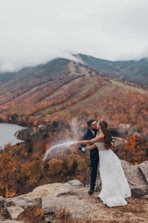 Wedding couple poses dramatically on mountain cliff with fall colors in valley below.