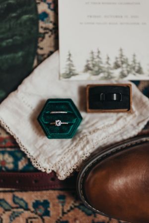 Close-up of wedding rings in teal velvet box with invitation and brown leather shoe details.