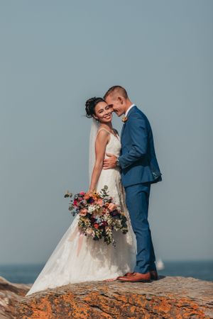 A couple in wedding attire shares an intimate moment on rocky coastal cliffs at sunset.