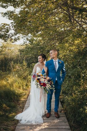 A couple in wedding attire walks along a wooden pathway surrounded by autumn foliage.
