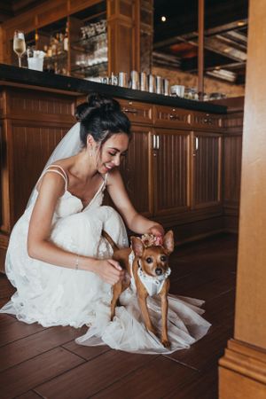 A person in a white wedding dress crouches down to pet a small dog on a wooden floor.