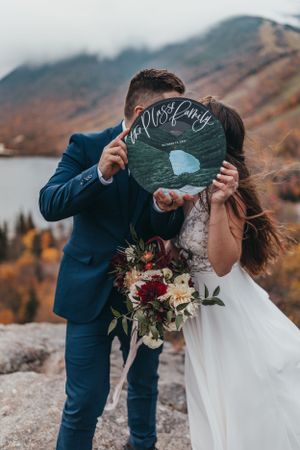Newlyweds hold up a chalkboard sign against a backdrop of fall foliage and mountains.