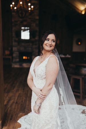 A bride in a detailed lace wedding gown poses indoors with soft lighting and wooden floors.