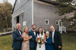 Wedding party poses for formal photos outside gray shingled building during fall celebration.