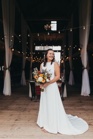 Bride in flowing white dress standing in rustic venue with string lights.