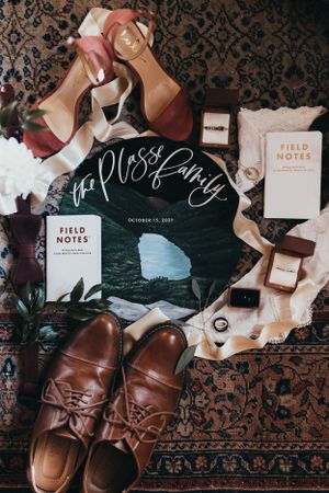 Wedding details flatlay with brown leather shoes, field notes, and decorative elements on dark pattern.