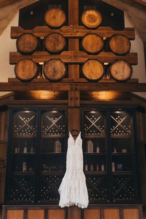 A white lace wedding dress hangs in front of wooden wine barrels and cabinets.