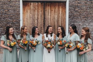 Bridesmaids in sage green dresses pose against a rustic brick wall with wooden doors holding vibrant fall bouquets.