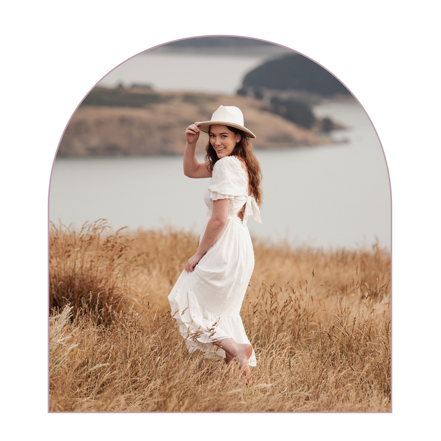 A romantic portrait in a golden field with a flowing white dress and sunhat against a misty lake backdrop.