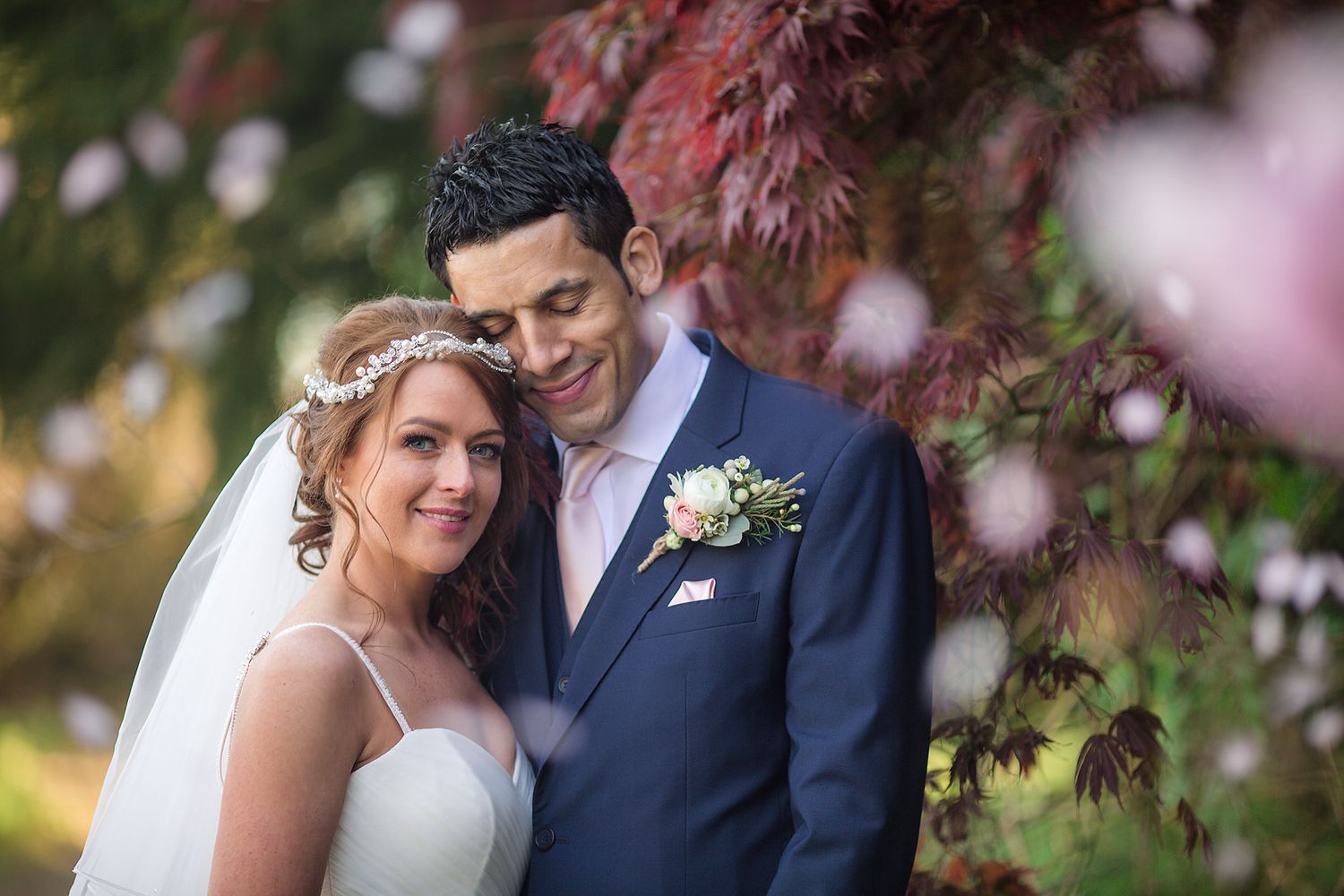 Bride and groom stand close together beneath a japanese acer tree with falling pink blossom