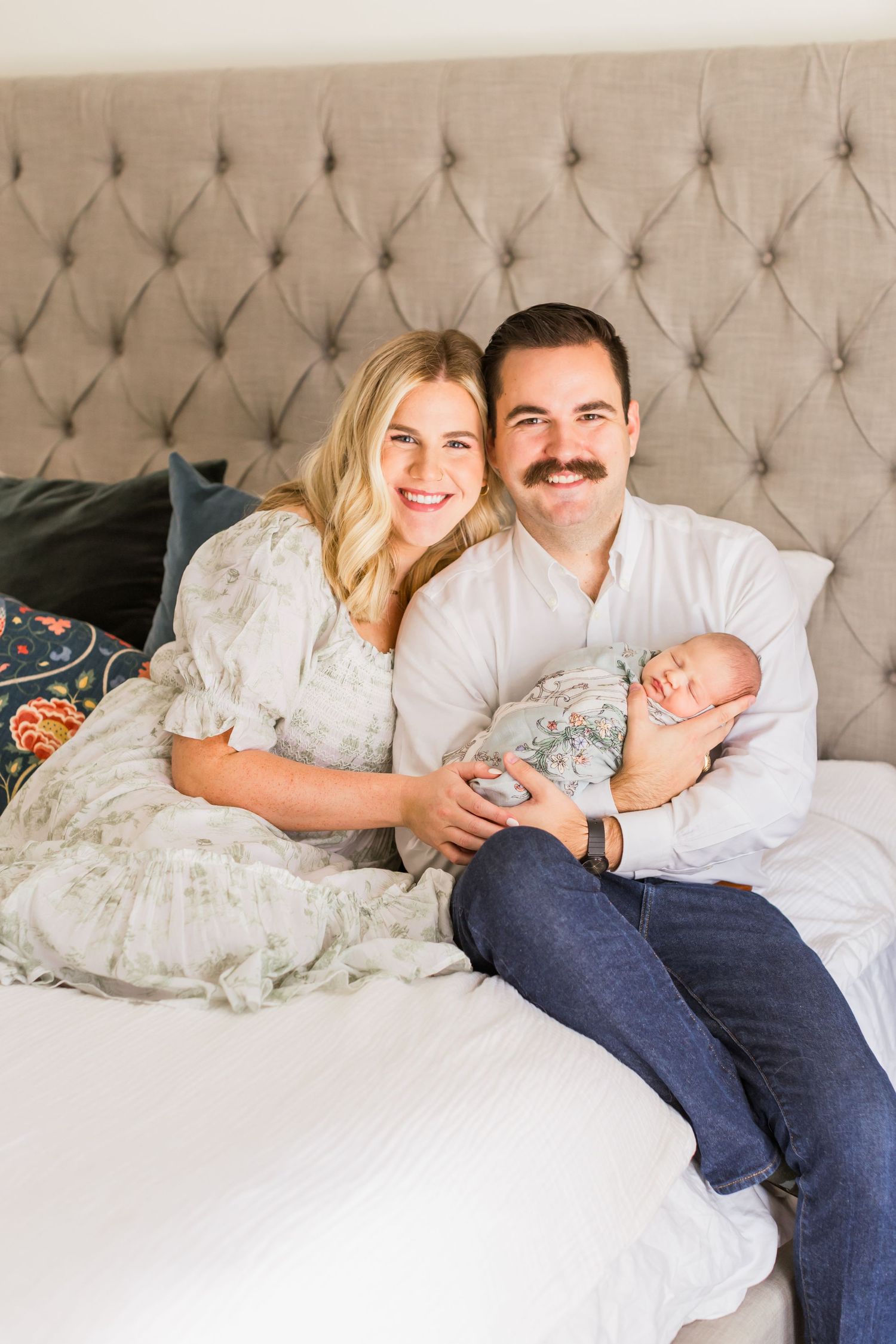 A family relaxes together on a tufted headboard bed with their newborn.