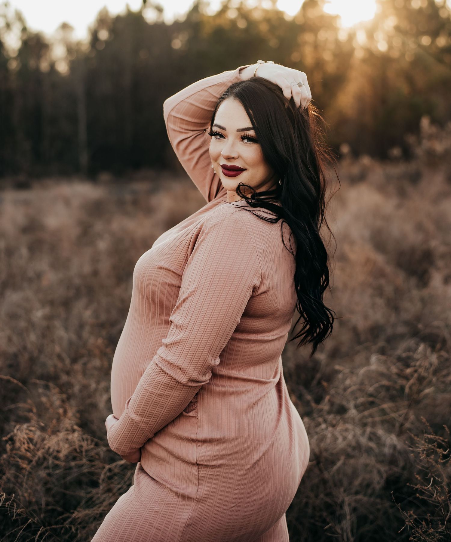 Pregnant woman in pink ribbed dress poses at golden sunset in an outdoor field.