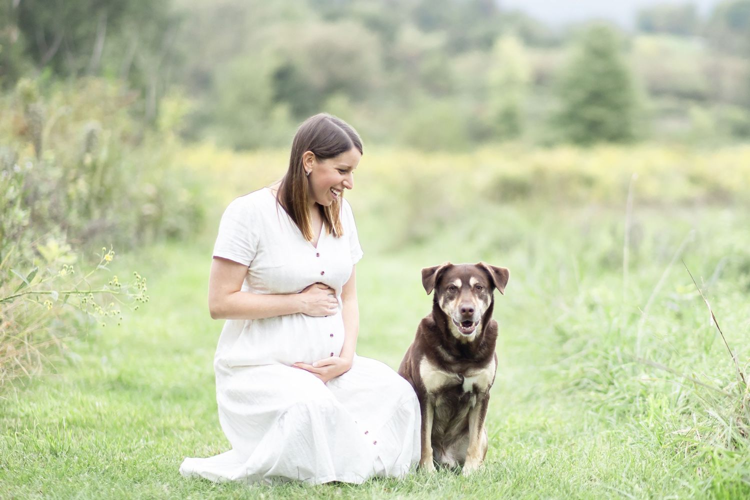 A woman in white dress sits with loyal dog companion in a serene meadow setting.