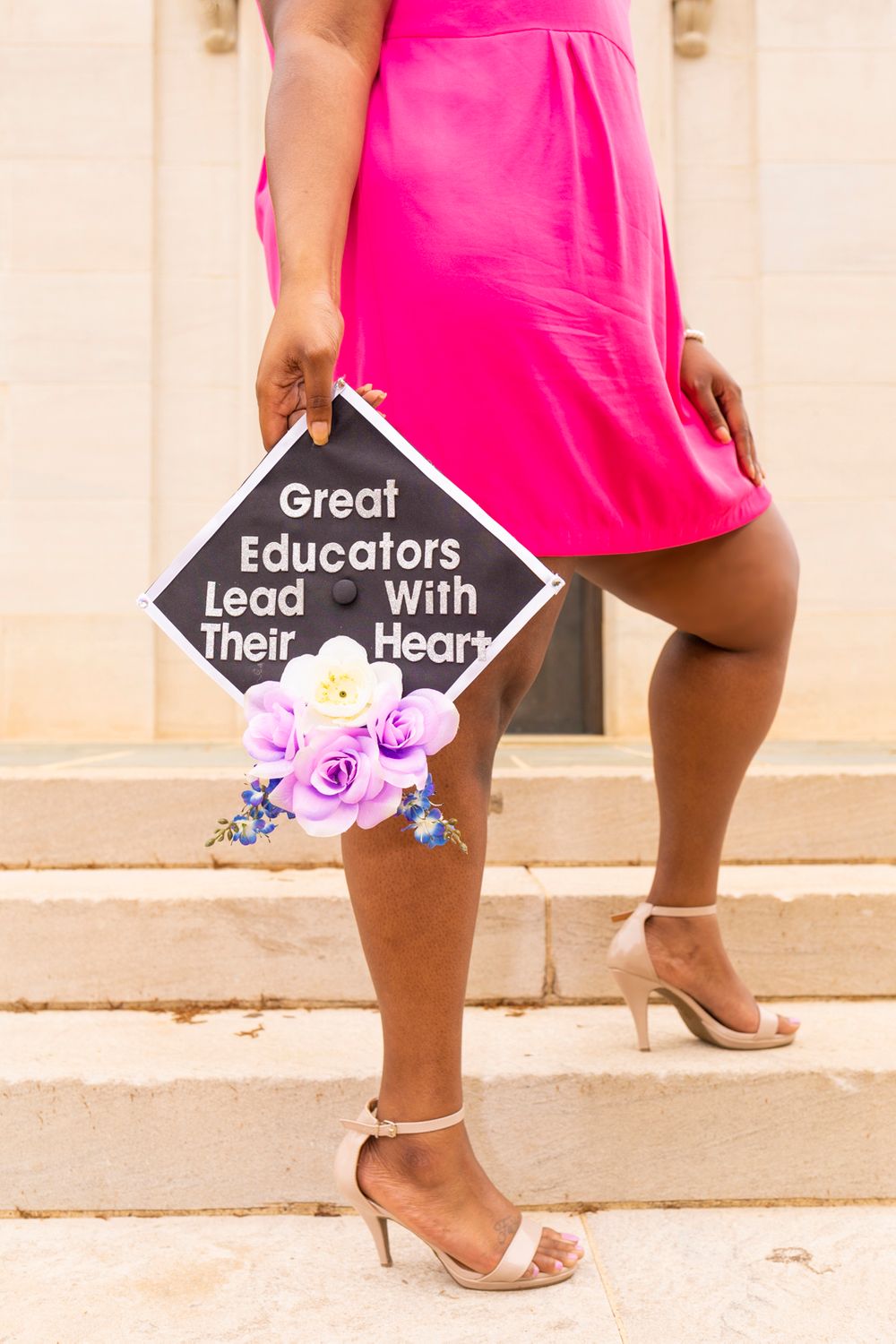 A grad standing on steps holding their grad cap with flowers that says "great educators lead with their heart."