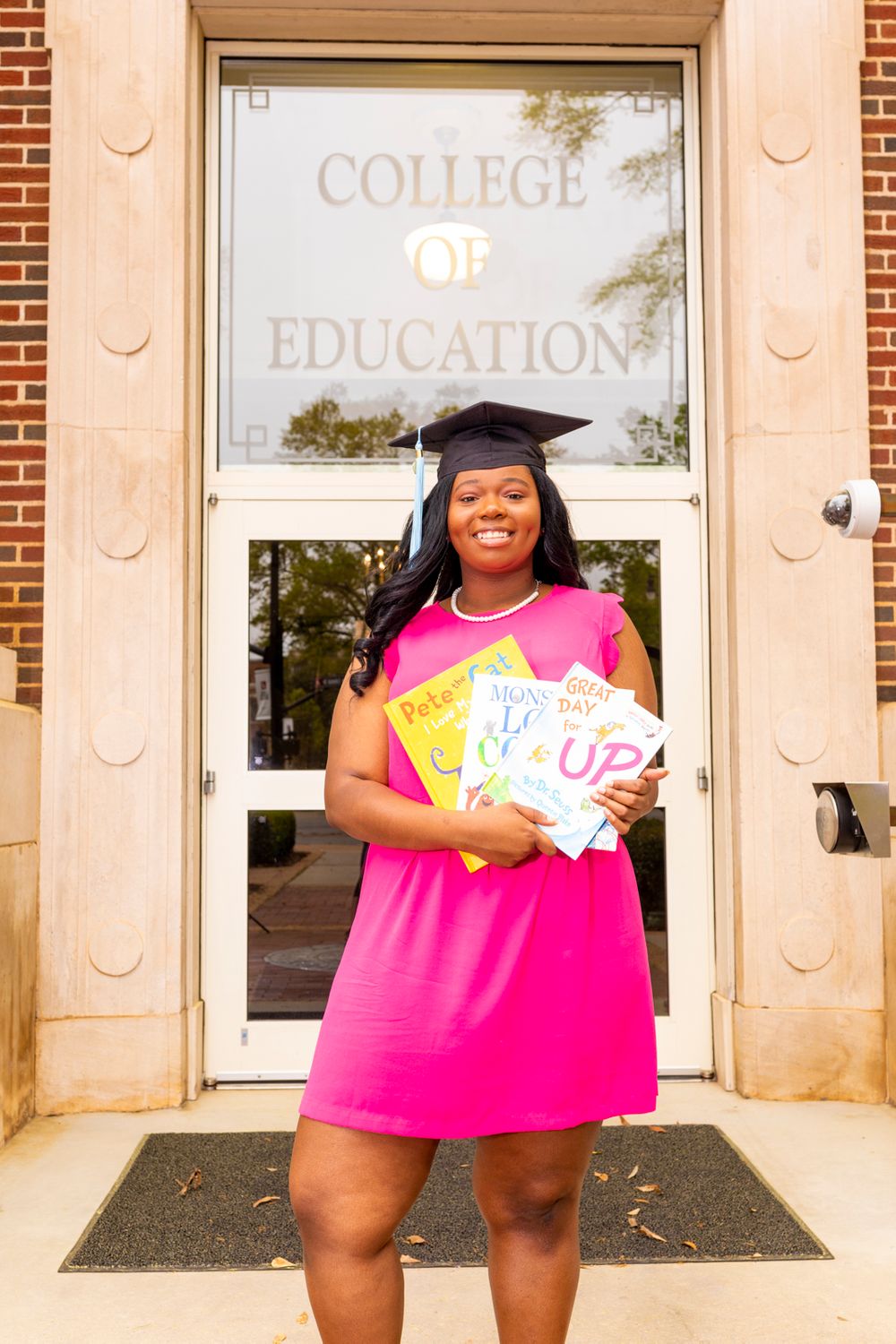 A grad wearing a hot pink dress standing in front of the "College of Education" holding three books.