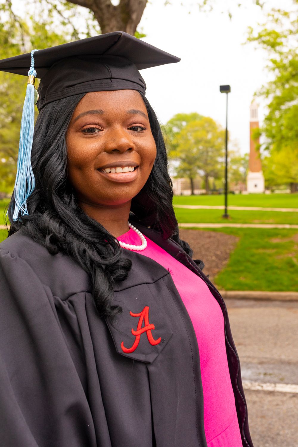 A grad smiling and wearing her grad gown over a hot pink dress standing with Denny Chimes in the background.