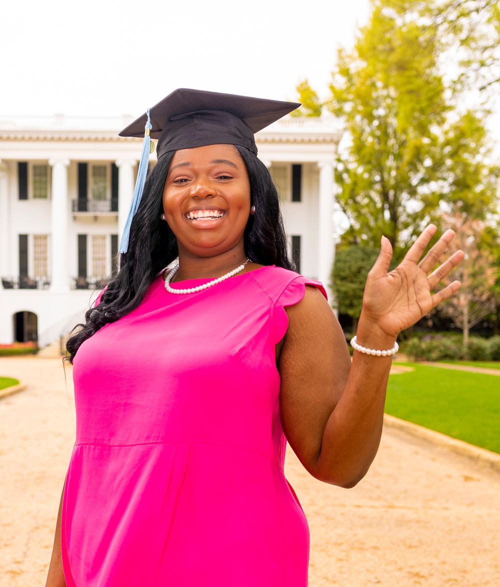A grad smiling and wearing a hot pink dress standing in front of the President's house waving.