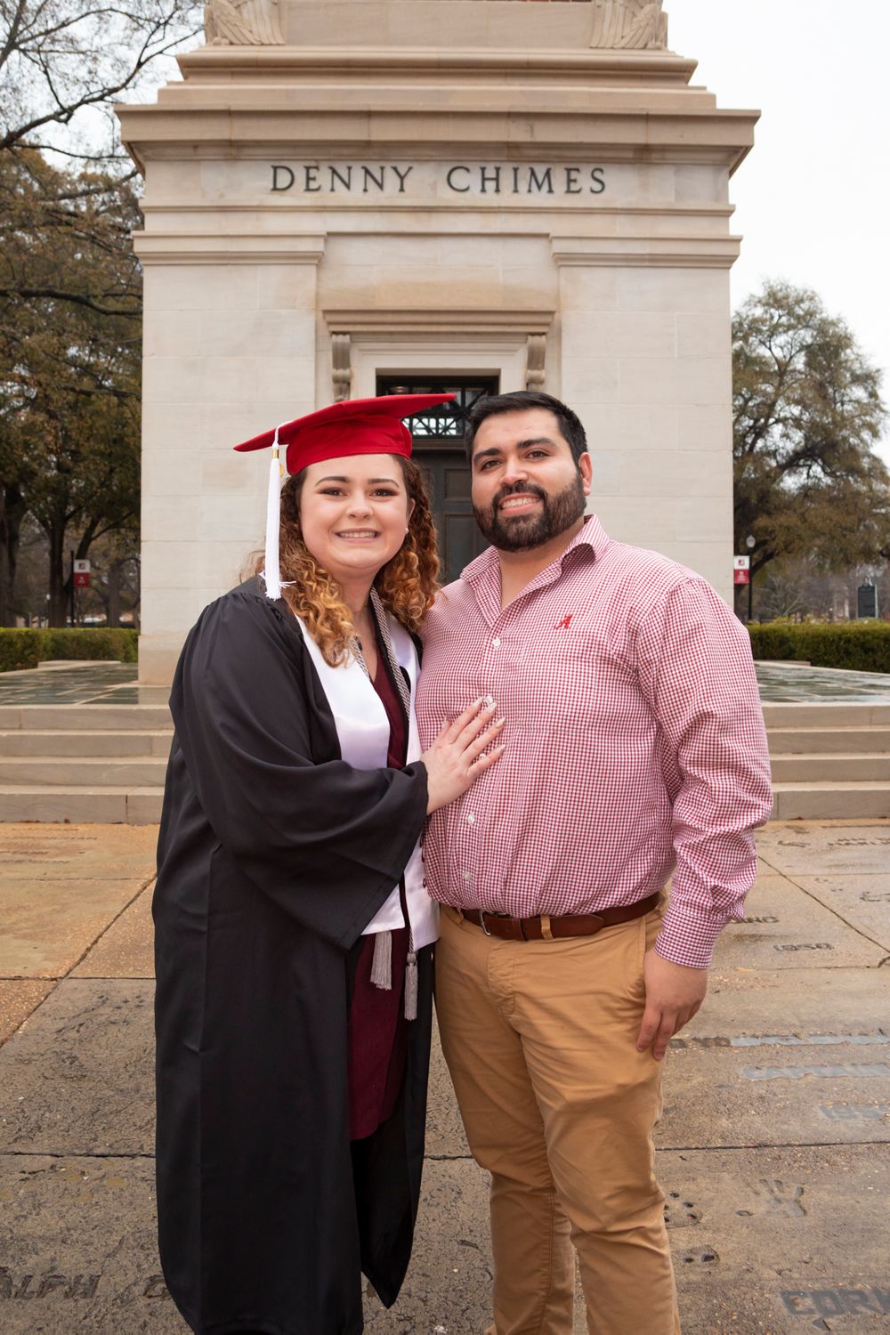 A grad wearing her grad gown, stole, tassel, and cap with her partner standing in front of Denny Chimes.