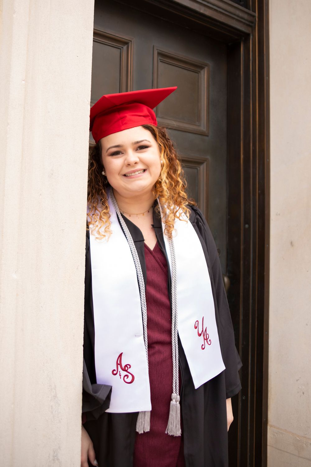 A grad wearing her grad gown, stole, tassel, and cap leaning on Denny Chimes door.