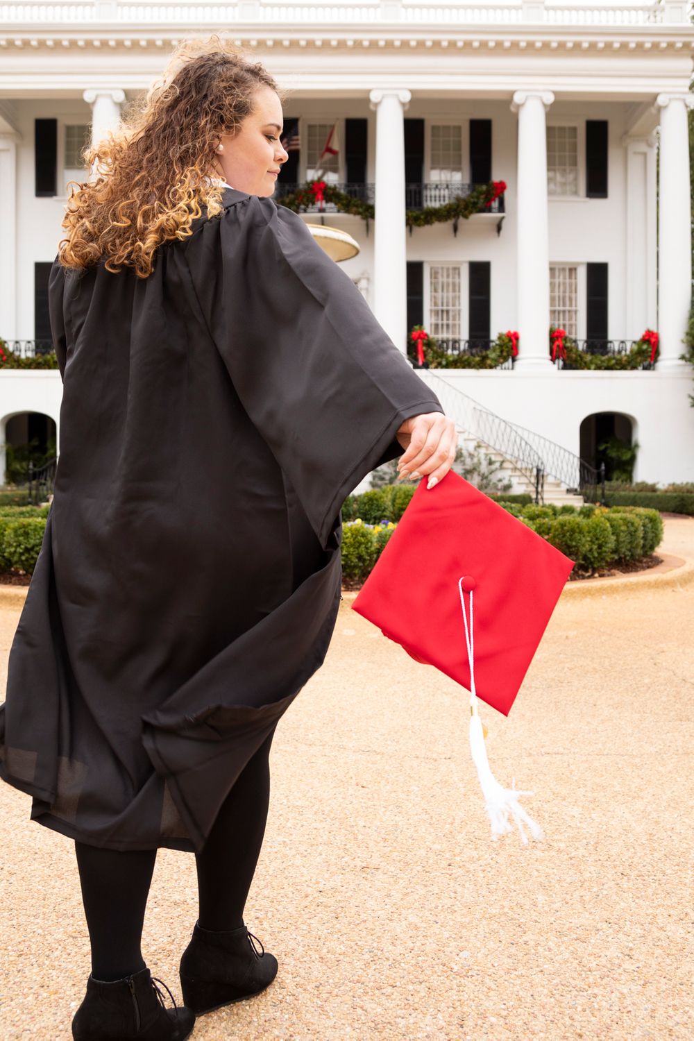 A grad wearing her grad gown, stole, and tassel walking towards the President house holding her red grad cap.