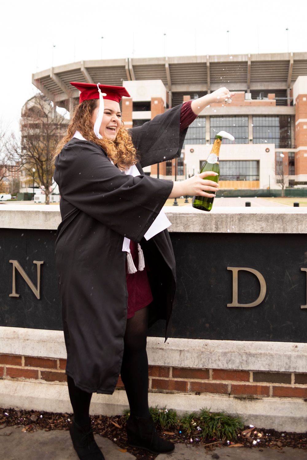 A grad wearing her grad gown, stole, tassel, and cap opening a bottle of champagne in front of the stadium..