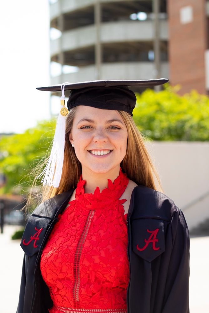 A grad wearing her a red dress with her grad gown and cap smiling at the camera.