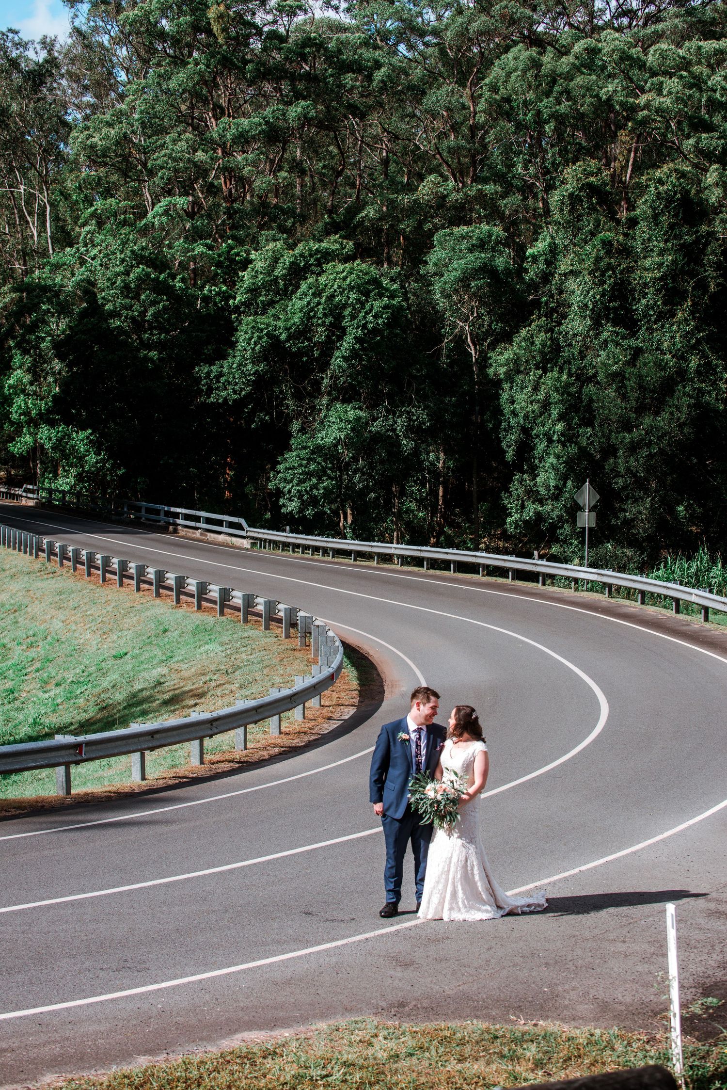 Newlyweds pose on a curved mountain road surrounded by tall trees.