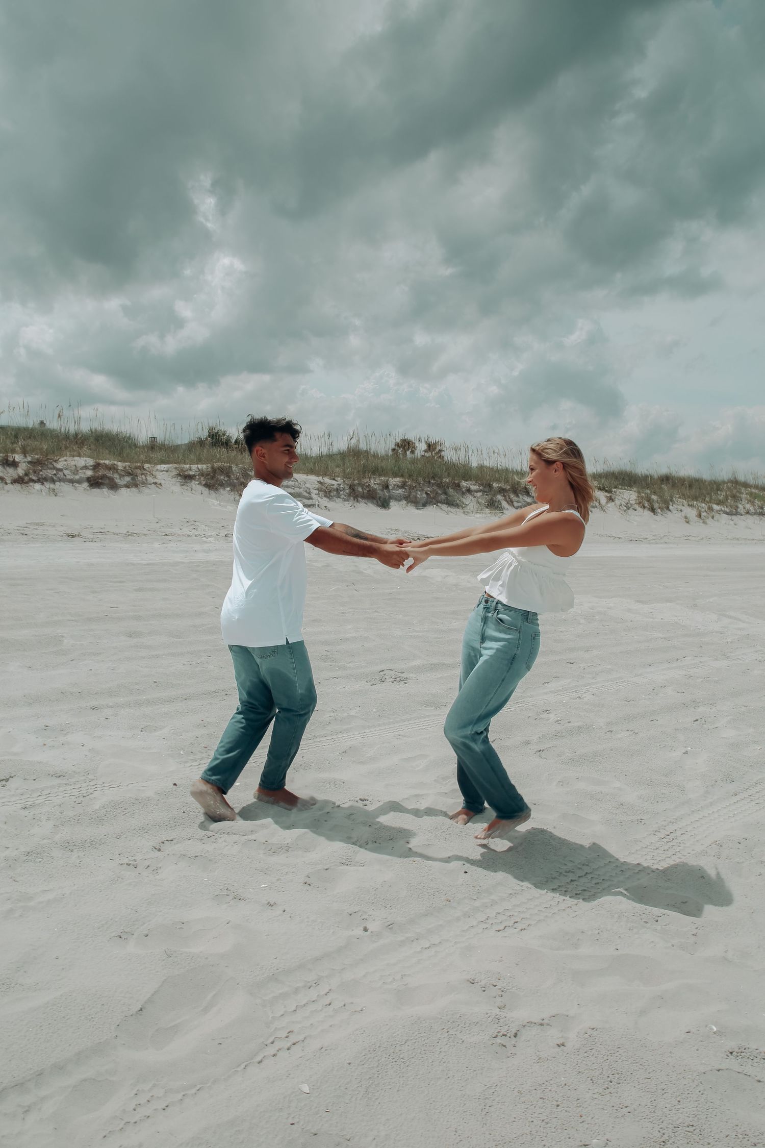 Happy couple holding hands and spinning together on a sandy beach under cloudy skies.